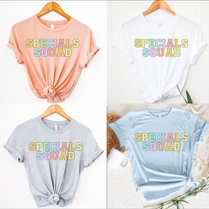 May include: Four t-shirts in different colors: peach, white, gray, and light blue. Each shirt has the words "SPECIALS SQUAD" printed in a colorful, block letter design. The shirts are displayed on wooden hangers.