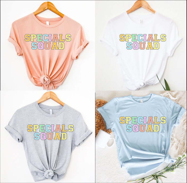 May include: Four t-shirts in different colors: peach, white, gray, and light blue. Each shirt has the words "SPECIALS SQUAD" printed in a colorful, block letter design. The shirts are displayed on wooden hangers.