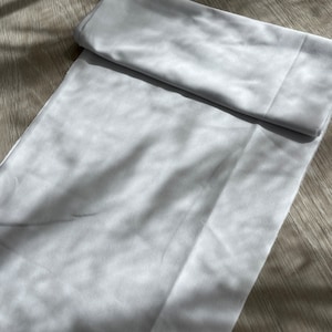 May include: A white fabric tablecloth with a smooth texture. The tablecloth is folded in half and lying on a wooden surface.