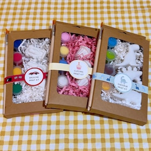 May include: Three rectangular brown cardboard boxes containing craft kits. Each kit includes white figurines, small paint pots, and a wooden paintbrush. The labels read Racecar Paint Kit, Cupcake Paint Kit, and Dinosaur Paint Kit with matching icons.
