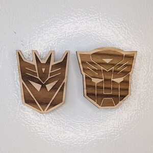 May include: Two wooden Transformers logos, one of Optimus Prime and one of Megatron, are displayed on a white surface. The logos are made of wood with a dark brown stain.
