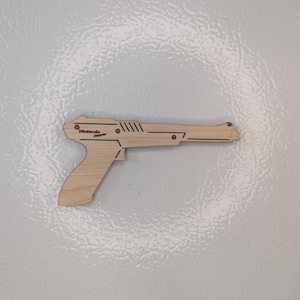 May include: A wooden Nintendo Zapper toy gun. The gun is light brown and has black lines and the text "Nintendo Zapper" printed on the side.