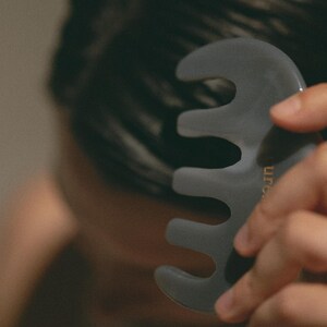 Puede incluir: Una herramienta de gua sha gris con cuatro puntas, utilizada para el masaje facial. La herramienta tiene la palabra "Urban" escrita en letras doradas.