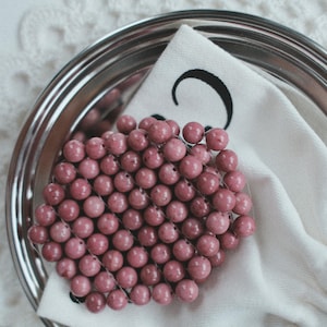 Op de afbeelding: Een cluster van roze kralen op een zilveren dienblad, rustend op een witte stoffen servet. De kralen zijn gerangschikt in een compacte, afgeronde vorm. De servet heeft een zwart ontwerp. Het dienblad staat tegen een witte, getextureerde achtergrond.
