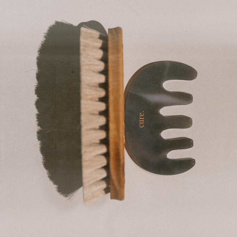 Puede incluir: Un cepillo de pelo de madera con cerdas negras y un cepillo de cerdas de jabal&iacute; blancas en un lado y un peine de pl&aacute;stico negro con la palabra "cure." en el otro.