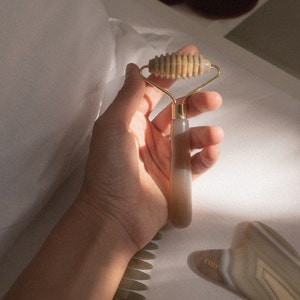 Puede incluir: Una mano sostiene un rodillo de jade con un mango dorado. El rodillo está hecho de una piedra marrón claro con una superficie texturizada. El rodillo se utiliza para el masaje facial.