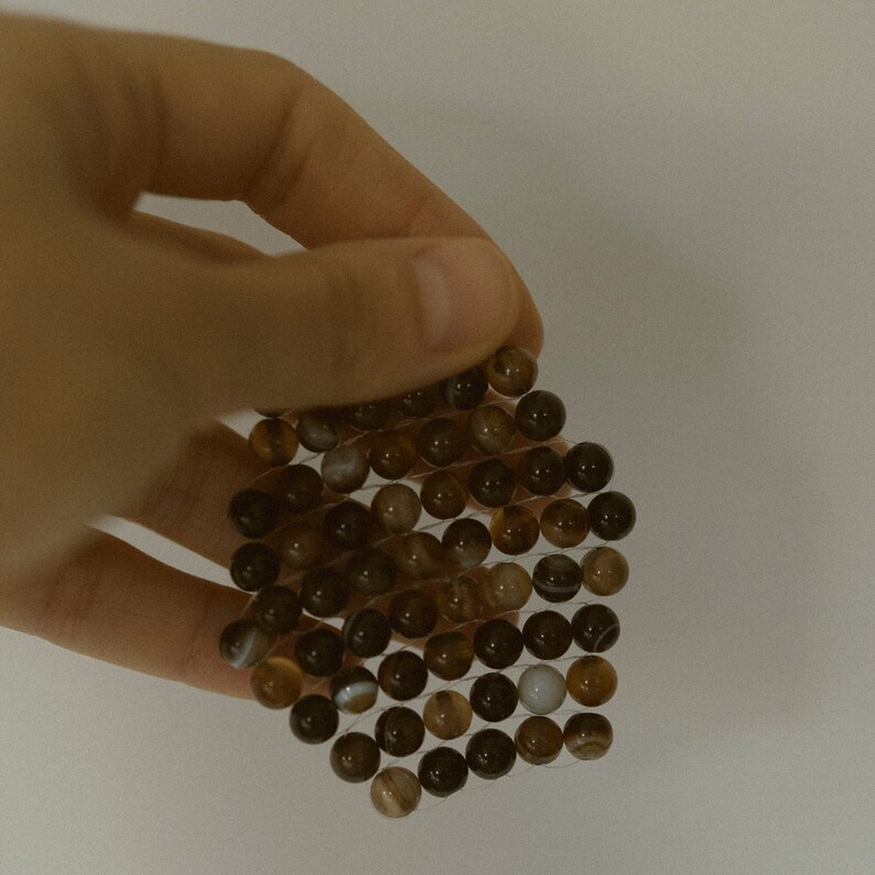 K&ouml;nnte beinhalten: Nahaufnahme einer Hand, die ein Armband aus braunen und wei&szlig;en Achatperlen h&auml;lt. Die Perlen sind in einem Gittermuster angeordnet und durch einen d&uuml;nnen Draht verbunden.