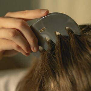 Puede incluir: Un masajeador de cuero cabelludo gris con tres puntas y la palabra "cure." impresa en el mango. El masajeador se est&aacute; utilizando en el cabello casta&ntilde;o de una persona.
