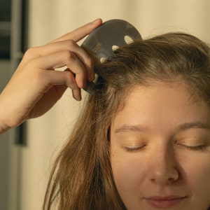 Puede incluir: Una persona con los ojos cerrados est&aacute; usando una herramienta de gua sha gris con la palabra "cure" para masajear su cuero cabelludo.