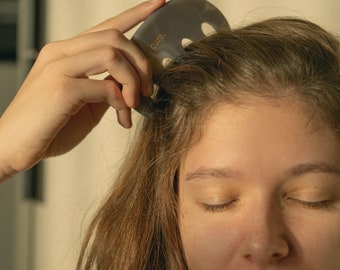 May include: A person with their eyes closed is using a gray gua sha tool with the word "cure" on it to massage their scalp.