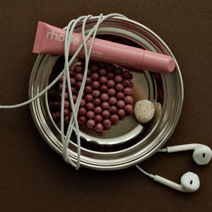 May include: A silver metal bowl containing a pink Rhode lip tint tube, a cluster of small dark red spherical beads, a white stone, and white wired earbuds wrapped around the items. The tube features the text "rhode" and "peptide lip tint".