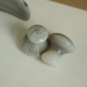 Op de afbeelding: Twee grijze en witte keramische gezichtsmassagetools. De bovenkant van een tool heeft een gouden inscriptie met de tekst "C."