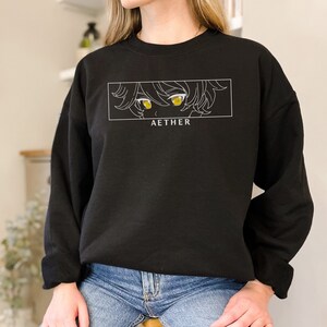 May include: Black sweatshirt featuring a white graphic of an anime character's eyes with yellow irises. The text "AETHER" is printed below the graphic.