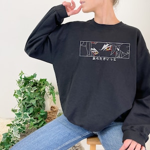 May include: Black sweatshirt with a white graphic of a character's eyes with red and yellow details. The text "あからた いって" is printed below the graphic.