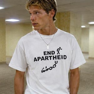 Puede incluir: Una camiseta blanca de manga corta con el texto negro END APARTHEID, un gr&aacute;fico de una figura de palo con mochila y una ilustraci&oacute;n de una cadena rota. La camiseta se combina con pantalones vaqueros azules y un cintur&oacute;n de cuero marr&oacute;n.