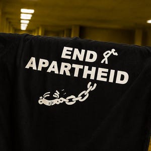 Puede incluir: Una camiseta negra de manga corta con el texto END APARTHEID impreso en blanco. El dise&ntilde;o incluye un gr&aacute;fico de una figura de palo y una cadena rota con un grillete, situados debajo de las letras may&uacute;sculas en negrita.