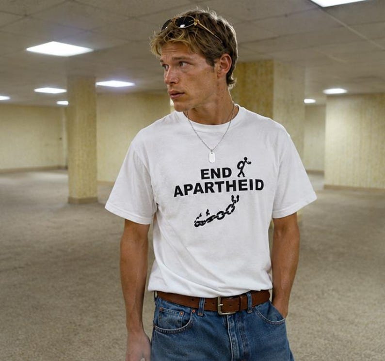 Puede incluir: Una camiseta blanca de manga corta con el texto negro END APARTHEID, un gr&aacute;fico de una figura de palo con mochila y una ilustraci&oacute;n de una cadena rota. La camiseta se combina con pantalones vaqueros azules y un cintur&oacute;n de cuero marr&oacute;n.