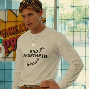 Könnte beinhalten: Ein weißes Langarmshirt mit dem schwarzen Schriftzug END APARTHEID, ergänzt durch eine Grafik eines Strichmännchens und zerbrochener Ketten. Das Oberteil wird mit einem beigen Ledergürtel und einer silberfarbenen Erkennungsmarke getragen.