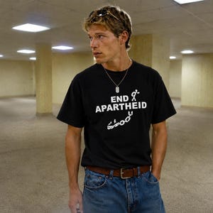 Puede incluir: Una camiseta negra de manga corta con el texto blanco END APARTHEID, acompa&ntilde;ado de un gr&aacute;fico de un monigote y cadenas rotas. La camiseta se lleva con vaqueros azules, un cintur&oacute;n de cuero marr&oacute;n y un collar con placa de identificaci&oacute;n.