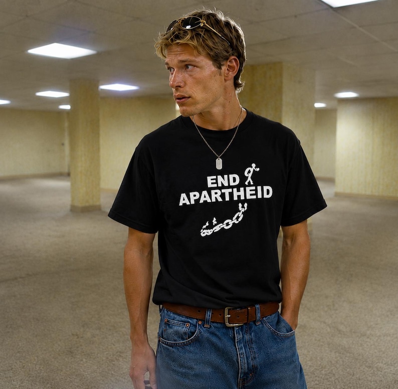 Puede incluir: Una camiseta negra de manga corta con el texto blanco END APARTHEID, acompa&ntilde;ado de un gr&aacute;fico de un monigote y cadenas rotas. La camiseta se lleva con vaqueros azules, un cintur&oacute;n de cuero marr&oacute;n y un collar con placa de identificaci&oacute;n.