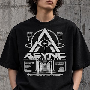 May include: A black short-sleeve t-shirt featuring a white graphic print. The design includes a large triangle with an eye, the text ASYNC RESEARCH DIVISION, and technical labels like SECTOR 04, AUTHORIZED ACCESS ONLY, and SOME PLACES ARE NOT MEANT TO BE FOUND.
