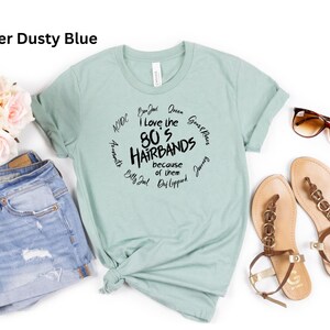 Peut inclure: Un t-shirt bleu poussi&eacute;reux avec le texte "I love the 80's Hairbands because of them" dans un design circulaire. Le t-shirt est pr&eacute;sent&eacute; avec un short en jean, des sandales et des lunettes de soleil. Un bouquet de fleurs roses est &eacute;galement pr&eacute;sent.