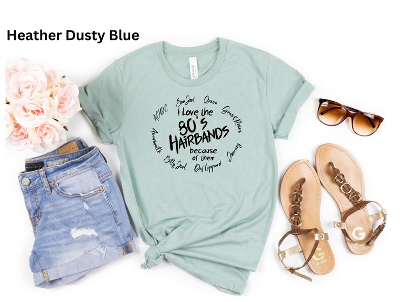 Peut inclure: Un t-shirt bleu poussi&eacute;reux avec le texte "I love the 80's Hairbands because of them" dans un design circulaire. Le t-shirt est pr&eacute;sent&eacute; avec un short en jean, des sandales et des lunettes de soleil. Un bouquet de fleurs roses est &eacute;galement pr&eacute;sent.
