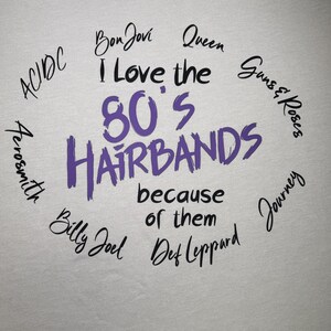 Peut inclure: T-shirt gris clair avec le texte "I Love the 80's Hairbands because of them" en violet et noir. Des noms de groupes comme Bon Jovi, Queen et Guns & Roses sont &eacute;galement pr&eacute;sents.