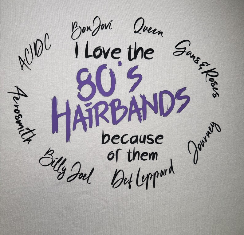 Peut inclure: T-shirt gris clair avec le texte "I Love the 80's Hairbands because of them" en violet et noir. Des noms de groupes comme Bon Jovi, Queen et Guns & Roses sont &eacute;galement pr&eacute;sents.