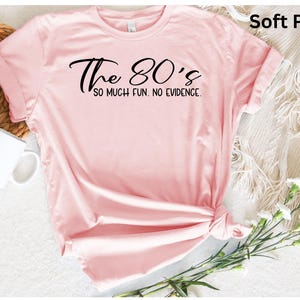 May include: Soft pink t-shirt with the text "The 80's so much fun. No evidence." in black script. The shirt is knotted at the bottom and has rolled-up sleeves. A white mug and flowers are in the background.