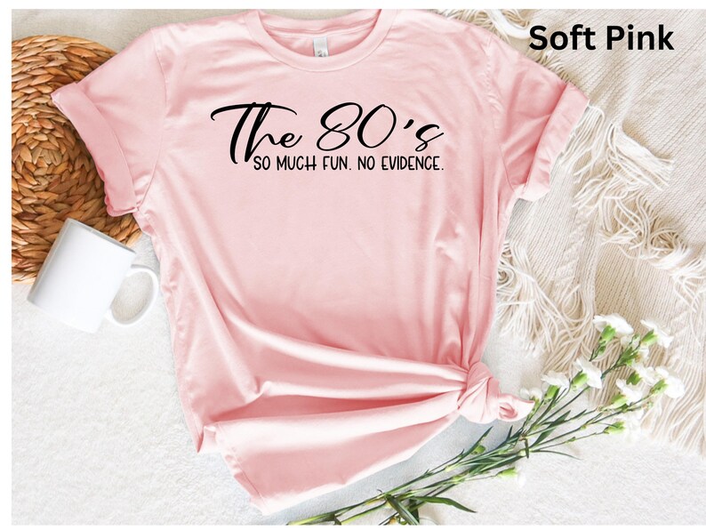 May include: Soft pink t-shirt with the text "The 80's so much fun. No evidence." in black script. The shirt is knotted at the bottom and has rolled-up sleeves. A white mug and flowers are in the background.