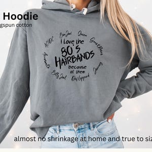 Peut inclure: Un sweat &agrave; capuche gris en 100% coton ringspun. Le devant affiche le texte "I Love the 80's Hairbands because of them" dans un design circulaire, avec des noms de groupes comme Bon Jovi et Def Leppard. Le texte "Grey Hoodie" est en haut.