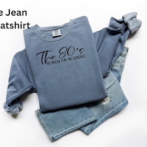 May include: A blue jean sweatshirt with the text "The 80's so much fun. no evidence" in black script. The sweatshirt is folded over a pair of blue jeans with distressed details. The clothing is on a white background.