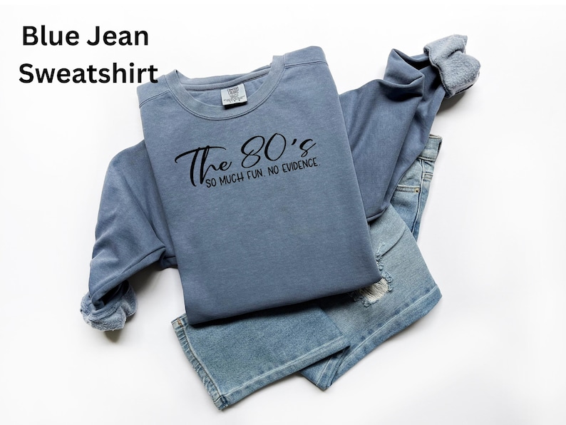 May include: A blue jean sweatshirt with the text "The 80's so much fun. no evidence" in black script. The sweatshirt is folded over a pair of blue jeans with distressed details. The clothing is on a white background.