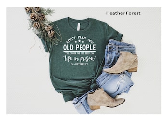 May include: Heather forest green t-shirt with the text "DON'T PISS OFF OLD PEOPLE" and "Life in prison is a deterrent". The shirt is paired with blue jeans and tan ankle boots. A sprig of pine is in the background.