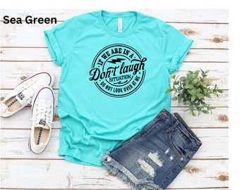 May include: Sea green t-shirt with rolled sleeves, featuring a black circular graphic that reads "If we are in a situation, Don't laugh, do not look over at me." White sneakers and denim shorts are also visible.