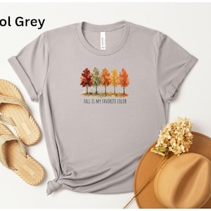May include: A cool grey short-sleeve t-shirt featuring a watercolor graphic of five trees with autumn-colored leaves in shades of red, orange, and yellow. Below the trees, the text reads "FALL IS MY FAVORITE COLOR" in a simple black font.