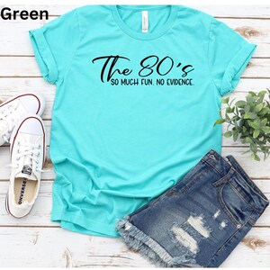 May include: Sea green t-shirt with the text "The 80's so much fun. no evidence." White sneakers and denim shorts are also in the image. The shirt is a casual style.