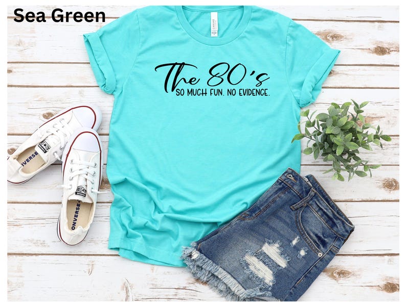 May include: Sea green t-shirt with the text "The 80's so much fun. no evidence." White sneakers and denim shorts are also in the image. The shirt is a casual style.