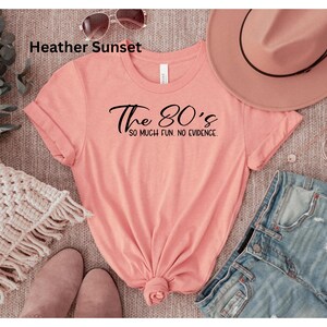 May include: A coral-colored t-shirt with the text "The 80's so much fun. no evidence" in black script. The shirt is styled with denim shorts, a pink hat, sunglasses, and shoes. A cozy blanket and greenery are also in the image.