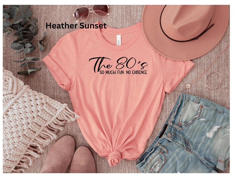 May include: A coral-colored t-shirt with the text "The 80's so much fun. no evidence" in black script. The shirt is styled with denim shorts, a pink hat, sunglasses, and shoes. A cozy blanket and greenery are also in the image.