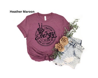 May include: Heather maroon t-shirt with the text "I match energy so how we gon' act today?" in a black, circular design. The design includes a peace sign made of a skeleton hand. The shirt is paired with denim shorts and decorative flowers.