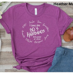 Peut inclure: T-shirt magenta avec l'inscription "I Love the 80's Hairbands" dans un motif circulaire blanc. Le t-shirt comprend des noms de groupes comme Bon Jovi et Def Leppard. Le t-shirt est associ&eacute; &agrave; un jean bleu et des sandales marron.