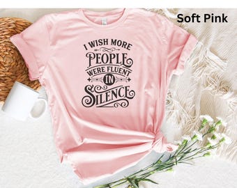 May include: Soft pink t-shirt with the text "I wish more people were fluent in silence" in black. The shirt is styled with rolled-up sleeves and a knot at the bottom. A white mug and flowers are in the background.