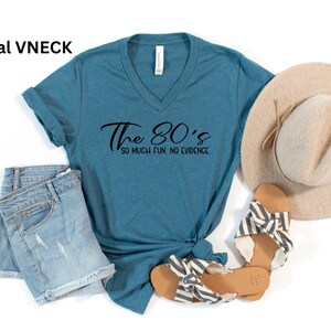 May include: Teal V-neck t-shirt with the text "The 80's so much fun, no evidence". The shirt is paired with denim shorts, a straw hat, and striped sandals. A casual, retro-themed outfit.