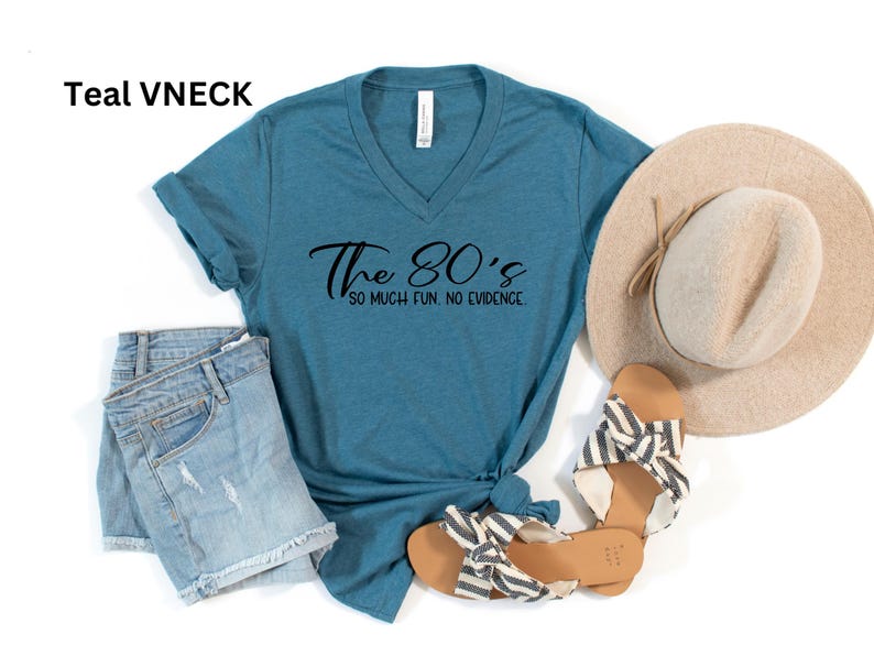 May include: Teal V-neck t-shirt with the text "The 80's so much fun, no evidence". The shirt is paired with denim shorts, a straw hat, and striped sandals. A casual, retro-themed outfit.
