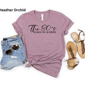 May include: A heather orchid t-shirt with the text "The 80's so much fun. no evidence." is paired with denim shorts, sunglasses, and gold-toned sandals. The casual outfit is laid out on a white surface.