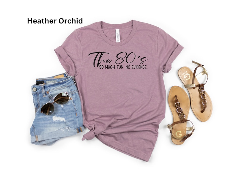 May include: A heather orchid t-shirt with the text "The 80's so much fun. no evidence." is paired with denim shorts, sunglasses, and gold-toned sandals. The casual outfit is laid out on a white surface.