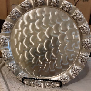 May include: A large, round, silver serving tray with a decorative, embossed border. The tray has a wavy, textured surface and a black metal stand.