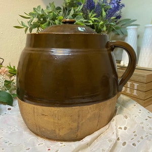 May include: A brown glazed ceramic crock with a lid and a handle. The crock has a brown glaze on the top half and a tan glaze on the bottom half. The crock is sitting on a white lace tablecloth.
